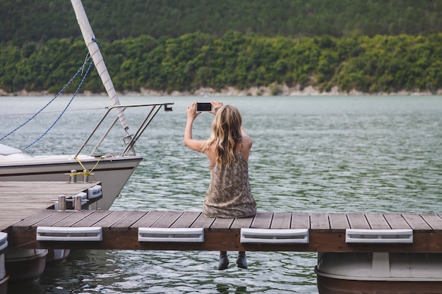 Girl blogger traveler sits on a pier and takes pictures on a smartphone