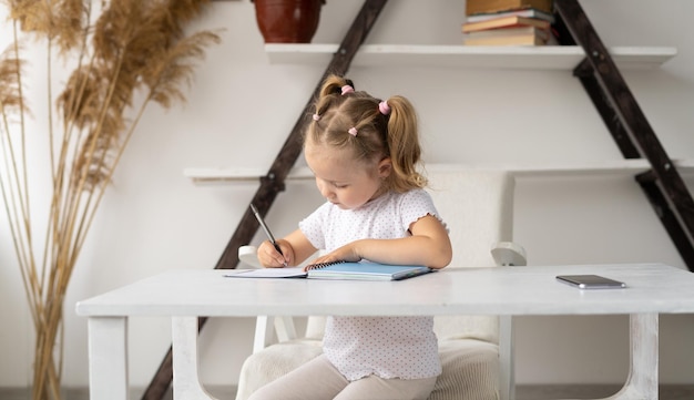 Girl blonde primary school student does homework at home at her desk Preschool child writes with a pen at home school baby learns to write handwritten in a notebook Distance education