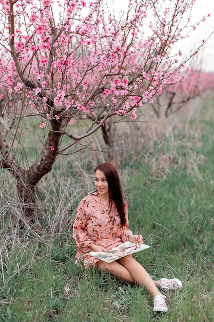 A girl in a blooming peach orchard is resting under a tree sitting and drawing