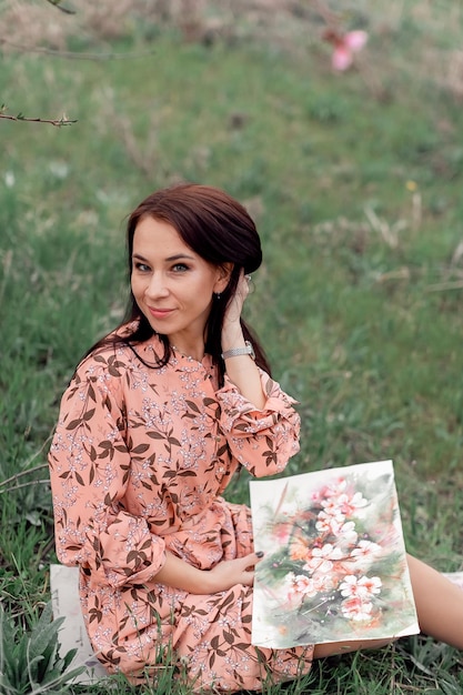 A girl in a blooming peach orchard is resting under a tree sitting and drawing