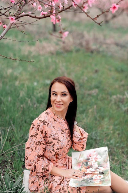 A girl in a blooming peach orchard is resting under a tree sitting and drawing