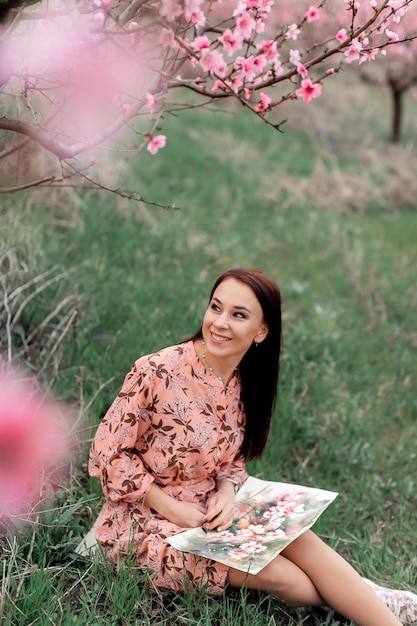 A girl in a blooming peach orchard is resting under a tree sitting and drawing