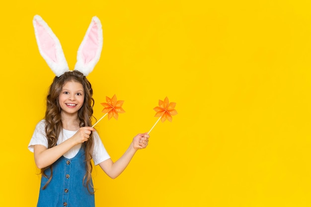 A girl in a blue dress with bunny ears holds a wooden stick and leaves on a yellow background.