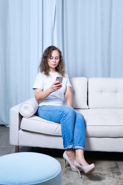 A girl in blue jeans and a white tshirt looks at the phone while sitting on a lightcolored sofa