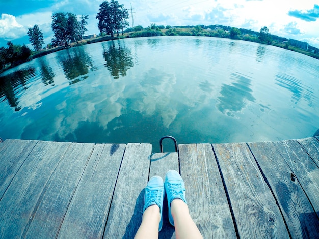 A girl in blue shoes sits on wooden pier near the lake river