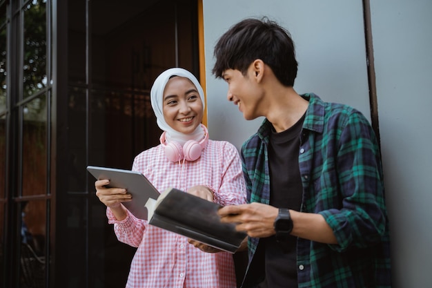 A girl and a boy using a digital tablet and read a book against a building wall in the background