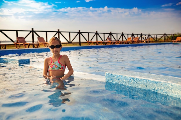 A girl in a bright swimsuit and swimming goggles poses near the pool sunbathing under the summer sun