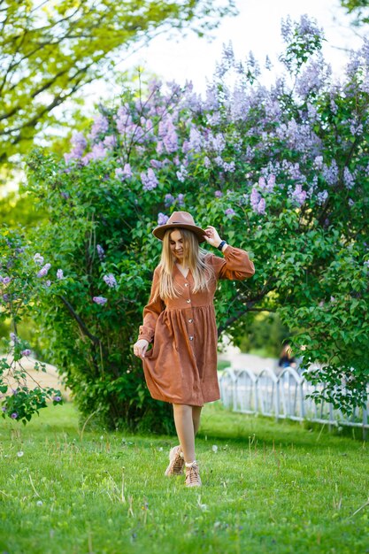 A girl in a brown hat and dress walks through the park, she has a European appearance and a smile on her face. Young attractive woman walks on a sunny day