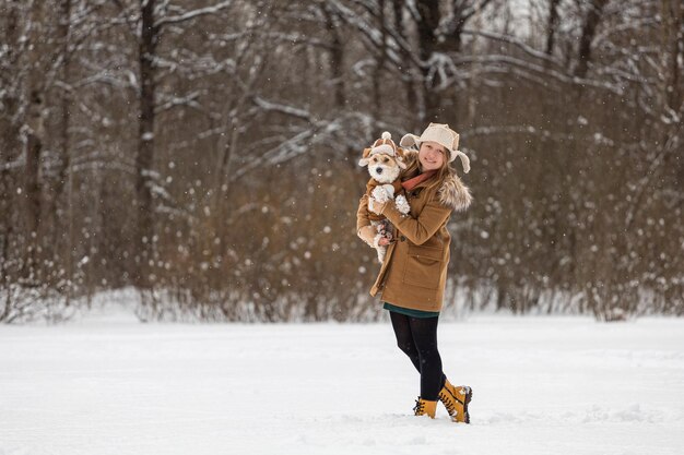 A girl in a brown hat with earflaps and a dog on the background of trees in the park Portrait of a Jack Russell Terrier dressed as a bear cub Snowing