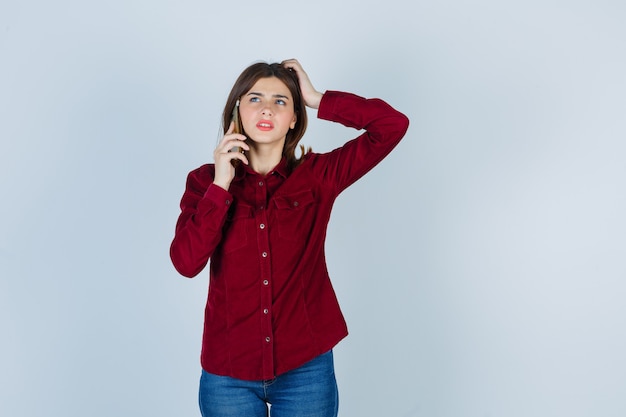 girl in burgundy shirt talking on smartphone, scratching head and looking thoughtful 