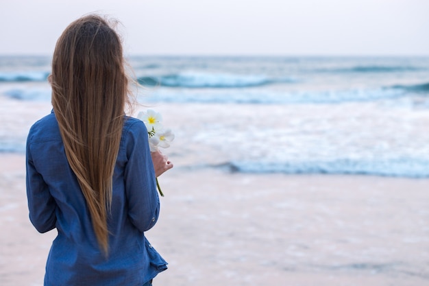 A girl by the ocean at sunset, holding a flower of plumeria in her hand