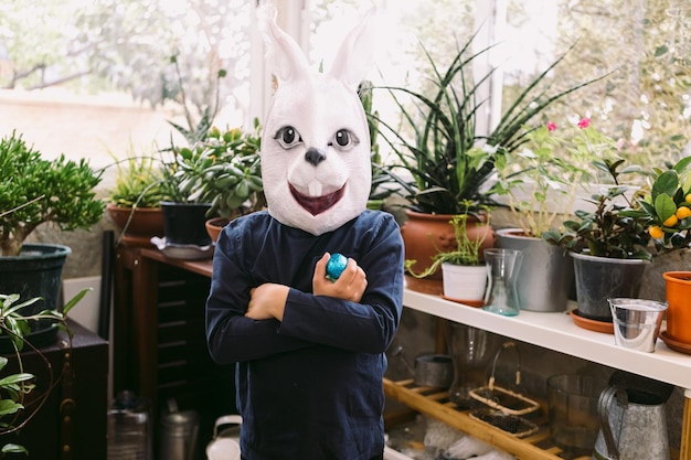 Girl celebrating Easter with a rabbit mask arms crossed and some chocolate eggs in a glassedin area surrounded by plants Easter celebration and costume concept