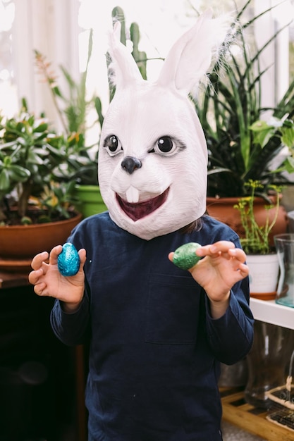 Girl celebrating Easter with a rabbit mask and some chocolate eggs in a glassedin area surrounded by plants Easter celebration and costume concept