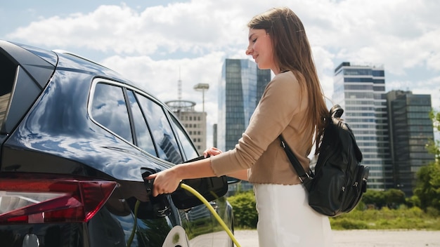 Girl charging her electric car at the parking lot near the city business center with skyscrapers