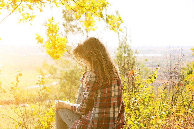 Girl in the checkered shirt is sitting in the autumn forest. Seasonal concept. Stylish hipster clothes outdoors. Nature philosophy around. Beautiful woman near the yellow leaves and trees.