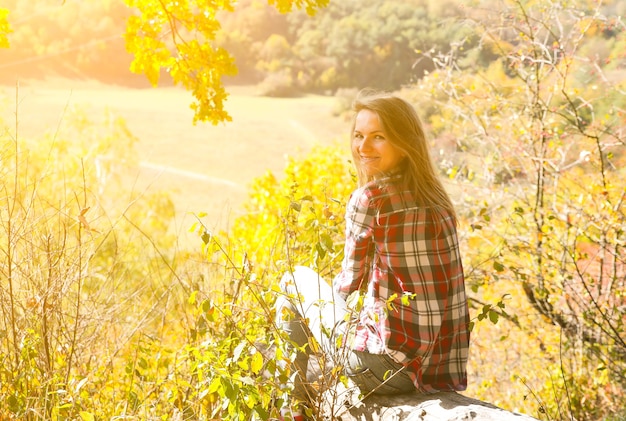 Girl in the checkered shirt is sitting in the autumn forest. Seasonal concept. Stylish hipster clothes outdoors. Nature philosophy around. Beautiful woman near the yellow leaves and trees.