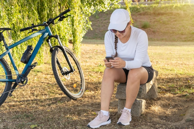 Girl checks her cell phone and rests under a tree next to her bike after exercising