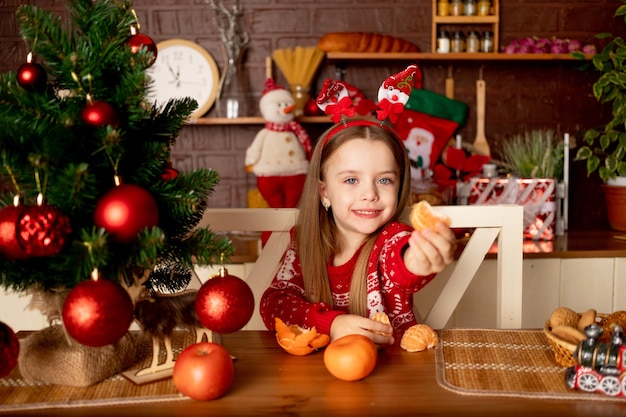 A girl child eats tangerines in a dark kitchen by a Christmas tree with red balls rejoices and smiles, the concept of new year and Christmas