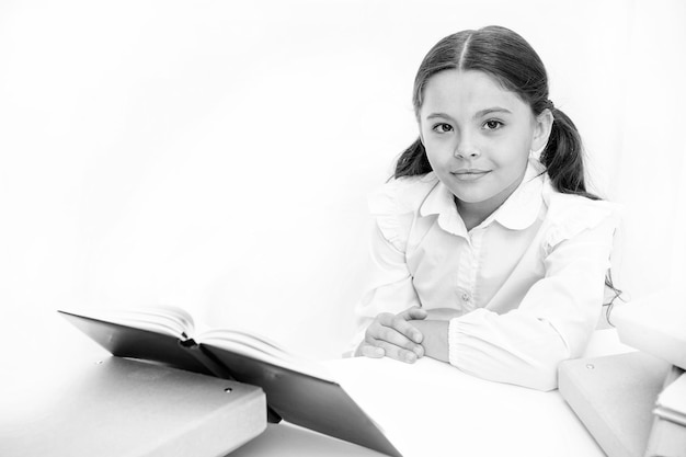 Girl child read book sit table white interior Schoolgirl studying textbook Kid school uniform happy face read book Excited about knowledge Homeschooling concept Interesting book for children