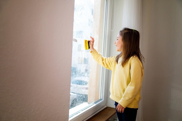 Girl child in yellow gloves with spray detergent with rag cleaning house washing windows springtime