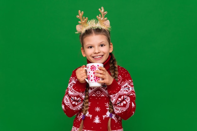 A girl in a Christmas sweater and reindeer horns drinks hot chocolate