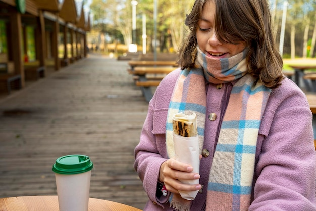 Girl in a coat eats street food standing at a table in an outdoor cafe