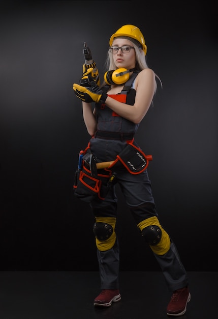 The girl in construction clothes and protective equipment posing with a screwdriver