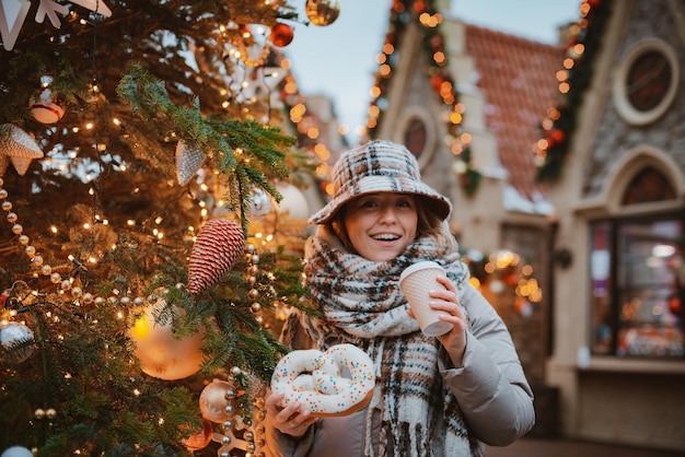 Girl cup hot drink while walking in Christmas market decorated with holiday lights in the evening. happy in big city.