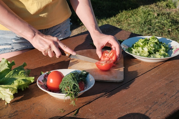 Girl cuts tomato on wooden board. Cooking in nature.