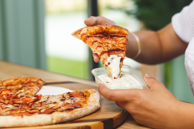 A girl dips a slice of pizza in sauce in a plastic container before eating it