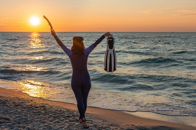 A girl diver on the beach at dawn raising her hands up