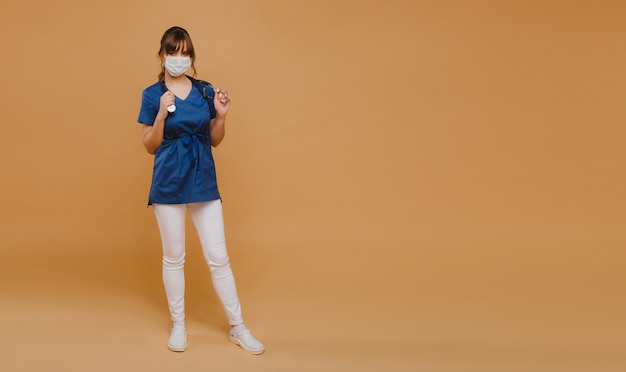 A girl doctor stands in a medical mask on a brown background and holds a stethoscope in her hands