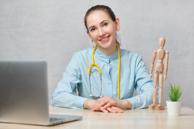 Girl doctor therapist sits at a table with a laptop on a white background and looks at the camera.