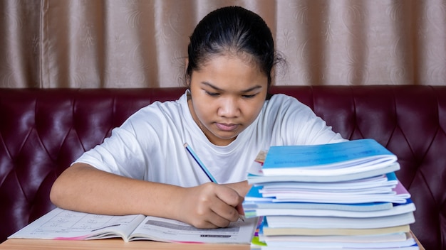 Girl doing homework on a wooden table and there was a pile of books next to it The background is a red sofa and cream curtains.