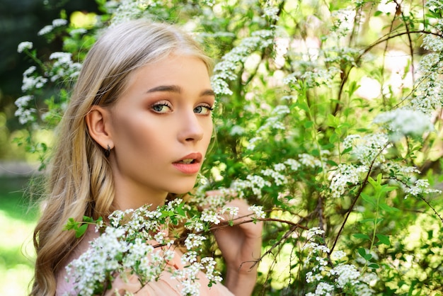 Girl on dreamy face, tender blonde near branches with white flowers. Young woman walks in park on sunny spring day