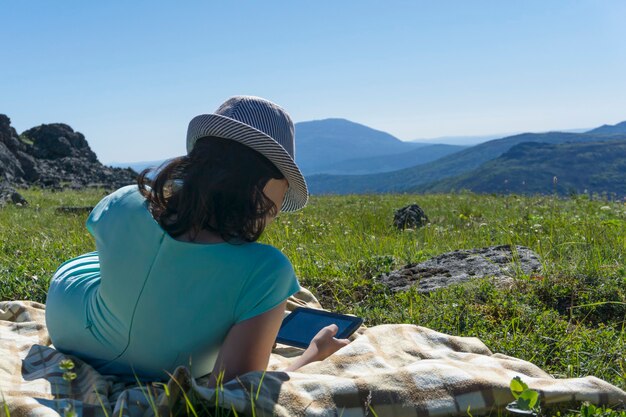 Girl in dress and hat sitting on a blanket with a tablet on an alpine meadow