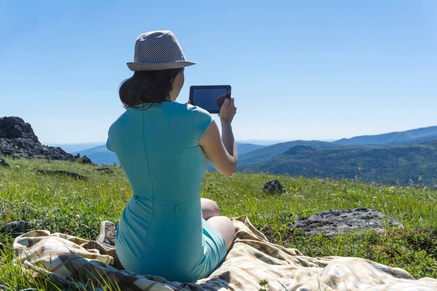 Girl in dress and hat sitting on a blanket with a tablet in her hands on an alpine meadow