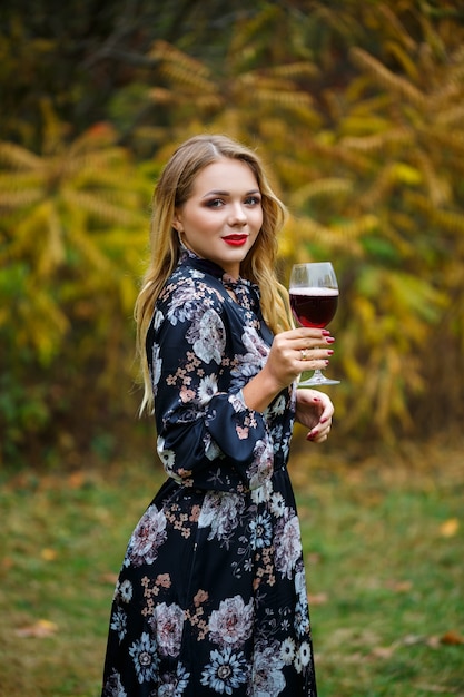 The girl in the dress holds a glass of red wine in the autumn forest