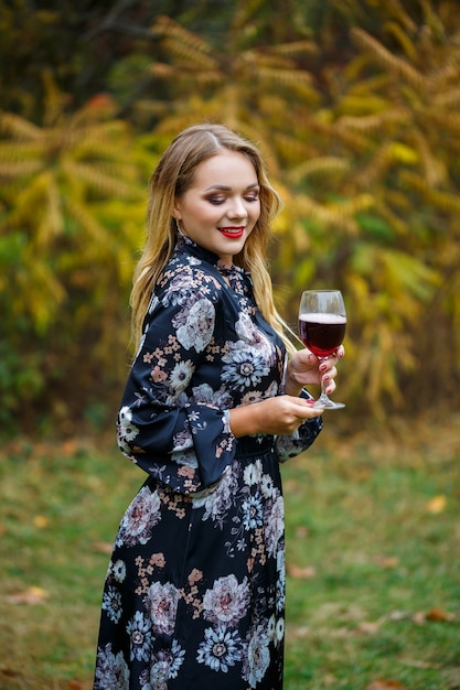 The girl in the dress holds a glass of red wine in the autumn forest