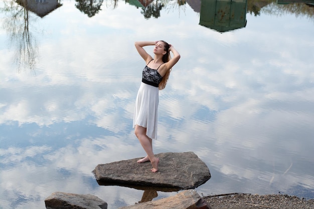 A girl in a dress on a stone in the river with a reflection of the sky with clouds in the water