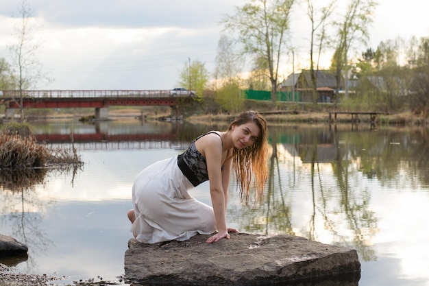 A girl in a dress on a stone in the river with a reflection of the sky with clouds in the water