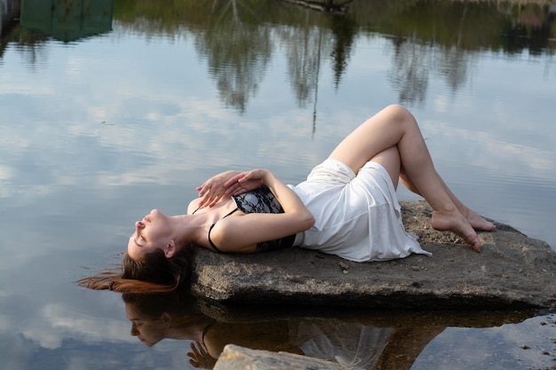 A girl in a dress on a stone in the river with a reflection of the sky with clouds in the water