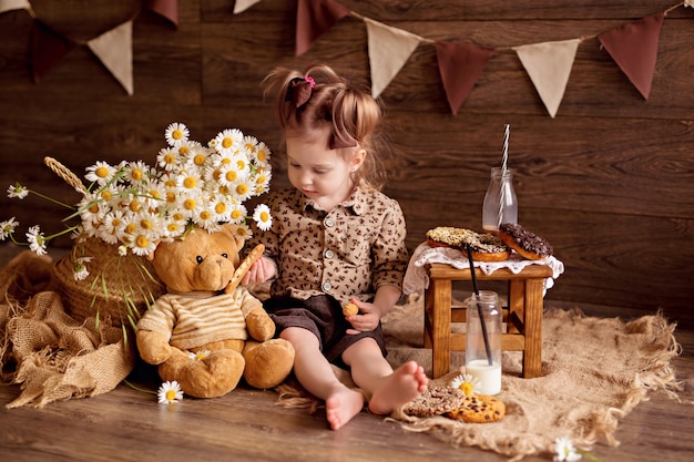 The girl eats cookies and feeds them with a teddy bear.