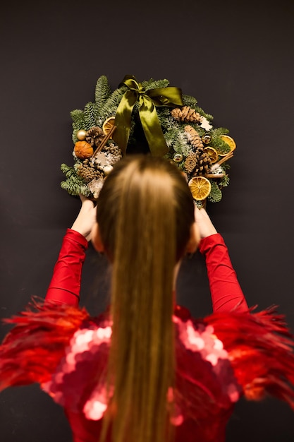 A girl in an elegant red dress hangs a Christmas wreath on the wall