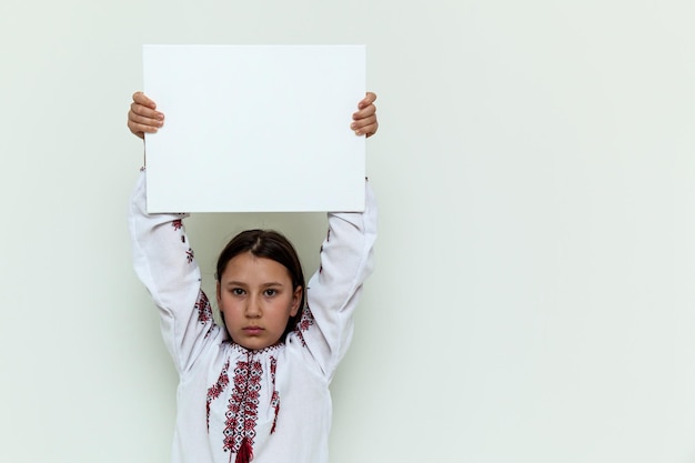 A girl in an embroidered dress stands against the background of a white wall holding a white sheet