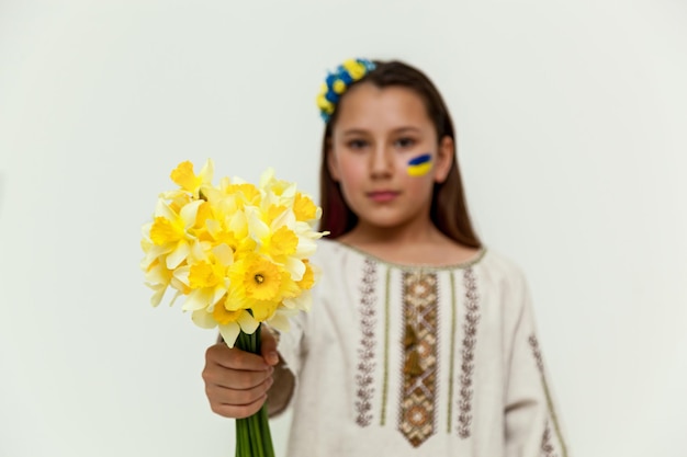 A girl in an embroidered dress stands against a white wall holding a bouquet of yellow daffodils