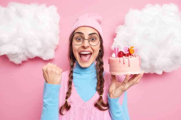 Photo girl exclaims loudly clenches fist from joy holds delicious cake celebrates birthday wears round spectacles pink hat casual clothes isolated on rosy 