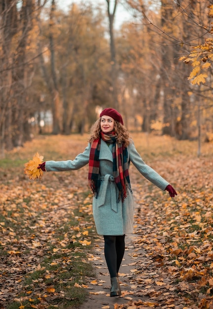 Girl on fabulous background of park with orange autumn leaves in hands walking on road. Pretty stylish smiling woman