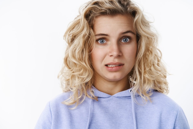 Girl feeling awkward and sorry as confessing in making mistake hum and haw with sorrow and reluctance standing intense and distressed, looking worried or nervous as posing in hoodie over white wall