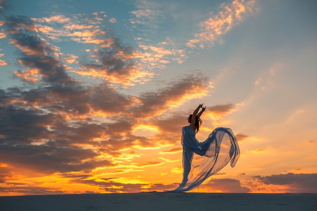 A girl in a fly white dress dances and poses in the sand desert at sunset